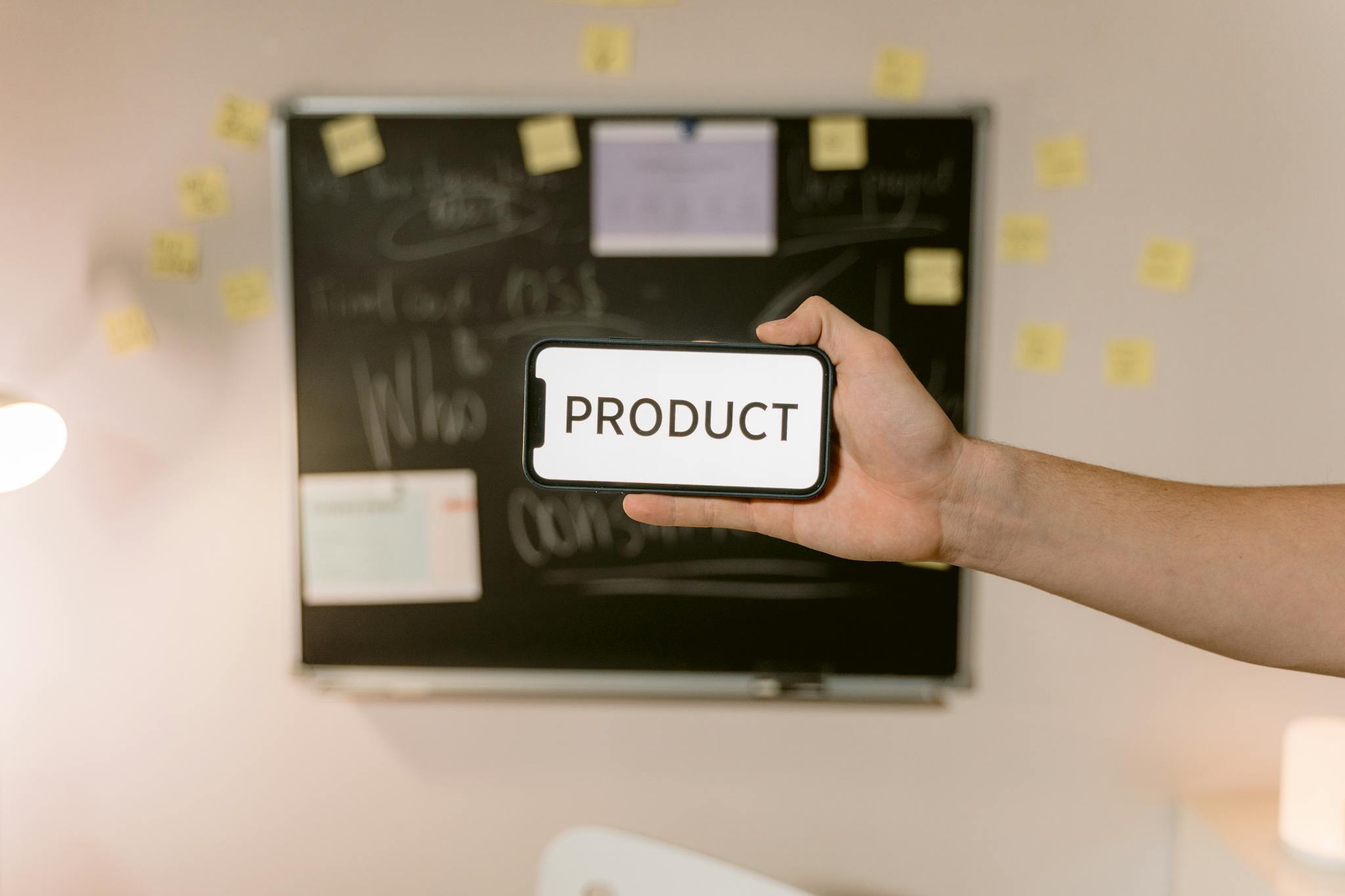 A hand holds a smartphone displaying the word 'PRODUCT' in front of a blurred office board.